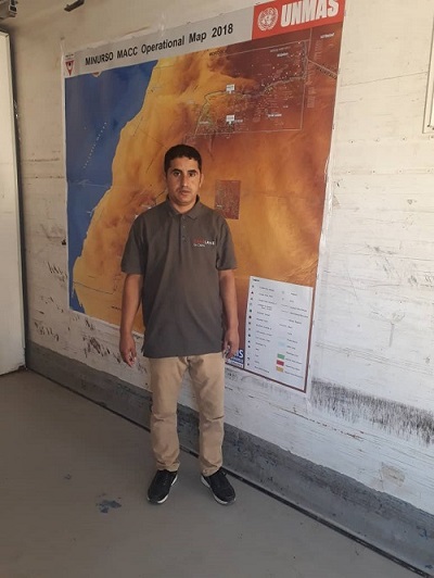 Abdallah Ammaisa - Deminer pictured in a dark grey SafeLane polo shirt and camel coloured trousers and black trainers. He is standing inside in front of a large map.