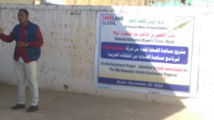 An image of one of leaders who supported the delivery of the the victim's assistance workshop in Sudan.  He is pictured to the left of the sign explaining that explains the workshop title in both Arabic and English. 